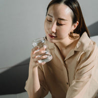 Load image into Gallery viewer, A lifestyle portrait of a model in a beige shirt holding a glass of water, reflecting a moment of hydration and wellness. This imagery aligns with the refreshing and replenishing benefits of the Calm Movements Eye & Cheek Mask, which uses hydrogel technology to soothe the skin.