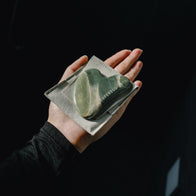 Load image into Gallery viewer, A hand holding the dark green marbled Nine-Dragon Jade Flow Egg Sculpting Gua Sha resting on its light gray microfiber pouch, showing the white SUPEREGG logo on both the tool and the pouch against a dark background.