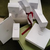 Load image into Gallery viewer, An overhead shot of the Full Ritual Set collection in their original white and olive green packaging. The arrangement includes the boxes for This Moment Hand Care, Sound Renewal Serum Cream, Vitamin Collective Vitality Elixir, and Calm Movements Eye & Cheek Mask, with a red gift ribbon tied around the Phyto Bounce box.