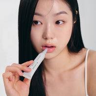 Load image into Gallery viewer, A close-up lifestyle shot of a model applying the Infinite Layers Lip Care. The model is using the white tube applicator to apply the nourishing lip treatment, showcasing its glossy finish and hydrating effect as part of the Full Ritual Set.