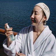 Load image into Gallery viewer, A person wearing a white bathrobe and the Plush Egg Spa Headband while spraying Kind Atmosphere Biome Mist on their face, set against a sunny ocean backdrop to evoke a refreshing self-care moment.