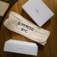 Load image into Gallery viewer, The Plush Egg Spa Headband laid out on a wooden surface between its minimalist white packaging boxes, one featuring the "BE A GOOD EGG" slogan and the other with product details.