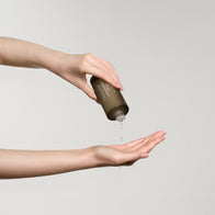 Load image into Gallery viewer, A lifestyle shot of a model's hands dispensing the Pure Transparency Treatment Essence. The dark olive bottle is held upside down, showing the clear liquid essence being poured into the palm, a key step in the Full Ritual Set routine.