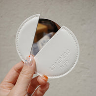 Load image into Gallery viewer, A hand holding the Reflection Egg Compact Mirror in its cream vegan leather case, showing the embossed "SUPEREGG NYC" logo and the stainless steel mirror's edge against a neutral wall.