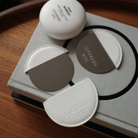 Load image into Gallery viewer, The Reflection Egg Compact Mirror with its cream-colored vegan leather case featuring an embossed SUPEREGG NYC logo, displayed on a book next to the This Moment Hand Care cream.