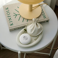 Load image into Gallery viewer, A lifestyle flat lay of the This Moment & Lucky Egg Duru Pouch set on a white side table. The spherical hand cream and beige drawstring pouch are arranged next to a Kinfolk book and a modern table lamp, creating a calm and mindful atmosphere.