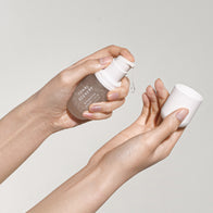 Load image into Gallery viewer, A close-up lifestyle shot of hands using the Tonal Scenery Reparative Serum. One hand holds the clear glass bottle while the other holds the cap, capturing the moment a clear droplet of the nutrient-rich serum is dispensed, emphasizing its fluid and hydrating texture.