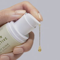Load image into Gallery viewer, A close-up shot of a hand with nude nail polish holding the Vitamin Collective Vitality Elixir by Superegg, with a single golden droplet of the serum being dispensed from the white pump against a soft grey background.