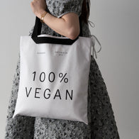 Load image into Gallery viewer, A lifestyle image of a model carrying the 100% Vegan Tote by its black handles. The white canvas bag features bold "100% VEGAN" typography and the Superegg logo, styled with a grey floral patterned dress for a modern, eco-conscious look.