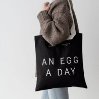 Load image into Gallery viewer, A lifestyle shot of the An Egg a Day Tote being worn over the shoulder. The image highlights the long white shoulder straps and the contrast of the black canvas bag against a model's grey knit sweater, emphasizing its practicality as an everyday carryall.