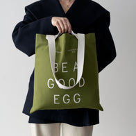 Load image into Gallery viewer, A lifestyle image of a model holding the Be a Good Egg Tote in olive green. The durable canvas bag features bold "BE A GOOD EGG" white typography and white twin carry handles, styled with a black coat for a modern, minimalist look.