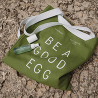 Load image into Gallery viewer, A flatlay lifestyle image of the Be a Good Egg Tote in olive green resting on a stone surface. The Kind Atmosphere Biome Mist is placed on the bag, highlighting the tote's bold white typography and its role as a perfect companion for skincare essentials.