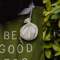Load image into Gallery viewer, A detailed lifestyle shot of the Be a Good Egg Tote in olive green with the Minimal Egg Cream Pouch hanging from its handle. The image highlights the contrast between the olive canvas and the cream vegan leather, set against a natural green leaf background.