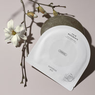 Load image into Gallery viewer, An artistic lifestyle shot of the Calm Movements Eye &amp; Cheek Mask sachet leaning against a smooth grey river stone. A delicate white magnolia branch with blooms accentuates the minimalist white packaging, reflecting the product's soothing and natural botanical ingredients.
