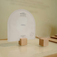 Load image into Gallery viewer, A minimalist display of the Calm Movements Eye &amp; Cheek Mask sachet and a clear acrylic mask-shaped cutout, both held upright by small wooden blocks. The warm, clean aesthetic emphasizes the unique patent-pending design of the hydrogel mask.