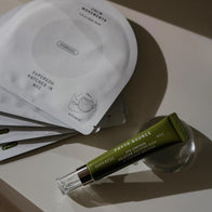 Load image into Gallery viewer, An aesthetic lifestyle shot of the Eye Care A Lot set. The Phyto Bounce Eye Contour Solution tube rests on a clear glass orb, placed atop several Calm Movements Eye &amp; Cheek Mask sachets. The composition features dramatic shadows and soft lighting to highlight the restorative eye care duo.