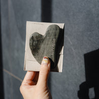 Load image into Gallery viewer, A hand with decorated nails holding the dark green marbled Nine-Dragon Jade Flow Egg Sculpting Gua Sha against its light gray microfiber pouch, showing the vertical SUPEREGG logo under bright, natural sunlight.
