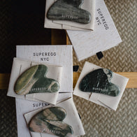 Load image into Gallery viewer, An overhead collection of multiple dark green marbled Nine-Dragon Jade Flow Egg Sculpting Gua Sha tools, each resting on a light gray protective pouch with the SUPEREGG NYC logo visible on both the jade stones and the fabric.