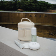 Load image into Gallery viewer, A lifestyle shot of the Superegg "In My Bag" set essentials—including the cream-colored vegan leather Little Egg Vanity Bag, facial mist, hand cream, and lip balm—arranged on a stone surface in an outdoor park setting.