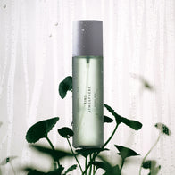 Load image into Gallery viewer, A refreshing lifestyle shot of the Kind Atmosphere Biome Mist standing behind green botanical leaves. The background features a glass pane with water droplets, emphasizing the mist's hydrating and skin-soothing properties.
