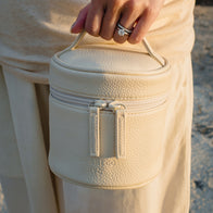 Load image into Gallery viewer, A close-up of a person wearing a diamond ring holding the Little Egg Vanity Bag in Eggshell cream by its top handle, showcasing the pebbled vegan leather and the debossed SUPEREGG NYC logo against a matching cream outfit.