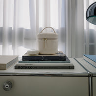 Load image into Gallery viewer, A lifestyle interior shot of the Little Egg Vanity Bag in Eggshell cream, placed on a stack of art books including "ROTHKO" on a modern white sideboard with a soft-focus window background.