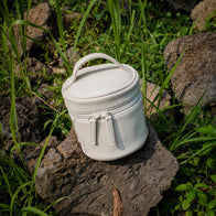 Load image into Gallery viewer, An outdoor lifestyle shot of the Superegg Little Egg Vanity Bag in Eggshell cream, resting on a dark volcanic stone surrounded by green grass, highlighting its pebbled vegan leather texture and debossed logo.