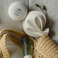 Load image into Gallery viewer, A lifestyle flat lay featuring the cream Lucky Egg Duru Pouch alongside the This Moment Hand Care and Kind Atmosphere Biome Mist. The products are tucked into a woven straw bag, emphasizing the pouch's role as a travel organizer for daily essentials.