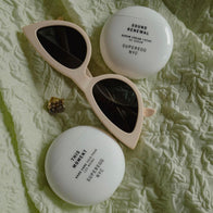 Load image into Gallery viewer, A stylized lifestyle flat lay of the New Yolker set. The Sound Renewal Serum Cream and This Moment Hand Care containers are nestled on a textured pale green fabric alongside a pair of chic, cream-colored cat-eye sunglasses.