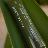 Load image into Gallery viewer, A vertical close-up shot of the Phyto Bounce Eye Contour Solution tube resting against a large, vibrant green leaf. The focus is on the sleek olive green packaging and the product name, highlighting its plant-based, restorative formula.