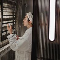 Load image into Gallery viewer, A side profile of a person in a white bathrobe and the Plush Egg Spa Headband, using a facial mist in a modern bathroom setting with elegant marble and glass textures.