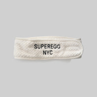 Load image into Gallery viewer, A cream-colored Plush Egg Spa Headband made of soft coral fleece, featuring an embroidered "SUPEREGG NYC" logo in black, shown against a clean gray background.