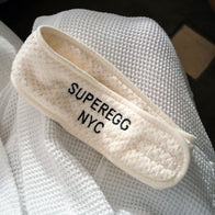 Load image into Gallery viewer, The cream-colored Plush Egg Spa Headband with an embroidered black SUPEREGG NYC logo, resting on a white waffle-weave robe or towel to highlight its soft, spa-like texture.