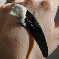 Load image into Gallery viewer, Close-up of the Superegg silver Precision Egg Skincare Spatula holding a dollop of white face cream against a person's hand, highlighting the engraved logo and smooth metallic finish.