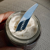 Load image into Gallery viewer, Close-up of the Superegg silver Precision Egg Skincare Spatula holding a dollop of white face cream against a person's hand, highlighting the engraved logo and smooth metallic finish.