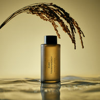 Load image into Gallery viewer, The Pure Transparency Treatment Essence bottle stands in a shallow pool of water with gentle ripples, beneath a golden stalk of grain, creating a serene and reflective aesthetic against a warm, soft-lit background.