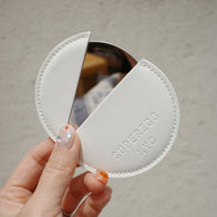 Load image into Gallery viewer, A hand holding the sleek, oval-shaped Superegg Reflection Egg Compact Mirror, showcasing its polished stainless steel surface and the engraved SUPEREGG logo against a clean, neutral background.