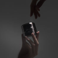 Load image into Gallery viewer, A cinematic shot of two hands interacting with the Soft Strength Rich Resilience Cream in a gunmetal silver jar, highlighting the reflective packaging and the SUPEREGG NYC logo against a moody, dark background.