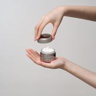 Load image into Gallery viewer, A pair of hands holding the Soft Strength Rich Resilience Cream in a gunmetal jar, with one hand lifting the lid to reveal the thick, white velvety cream inside against a clean gray background.