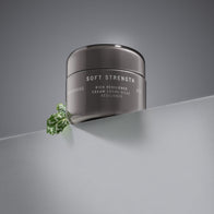 Load image into Gallery viewer, A sleek gunmetal jar of Superegg Soft Strength Rich Resilience Cream positioned on a gray slanted surface with a green leaf peeking from behind, showcasing the SUPEREGG NYC logo and "Crème Riche Résilience" text.