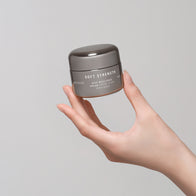 Load image into Gallery viewer, A lifestyle shot of a hand holding the Soft Strength Rich Resilience Cream jar against a minimalist grey background. The image showcases the sleek, pebble-shaped grey jar of this moisturizing cream, which is a core component of the Full Ritual Set.