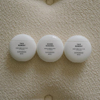 Load image into Gallery viewer, A top-down lifestyle shot of three minimalist white egg-shaped jars resting on a textured cream fabric. The Sound Renewal Serum Cream is centered between two jars of This Moment Hand Care, highlighting the cohesive and elegant design of the Superegg product line.
