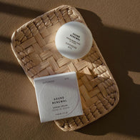 Load image into Gallery viewer, A warm, sun-drenched lifestyle shot of the Sound Renewal Serum Cream. Both the minimalist white egg-shaped jar and its square outer packaging are displayed on a woven straw mat, casting soft shadows that highlight the clean and elegant brand aesthetic.