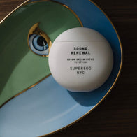 Load image into Gallery viewer, A top-down artistic shot of the Sound Renewal Serum Cream resting on a decorative ceramic plate with gold-rimmed teal and white sections. The minimalist white egg-shaped jar is highlighted against the elegant, curved lines of the plate, reflecting a sophisticated skincare lifestyle.