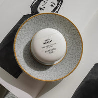 Load image into Gallery viewer, An artistic top-down lifestyle shot of the This Moment Hand Care egg-shaped container centered on a mosaic-patterned decorative plate. The setting includes a black napkin and a sketch of a face, reflecting the brand's intersection of beauty, art, and modern design.