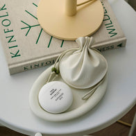 Load image into Gallery viewer, A top-down aesthetic shot of the This Moment Hand Care egg-shaped container resting on a curved cream-colored tray. Beside it is a matching drawstring pouch and a Kinfolk book, creating a serene, minimalist lifestyle scene that emphasizes the product's portable and elegant design.