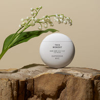 Load image into Gallery viewer, The spherical white This Moment hand cream container is placed on a rugged, natural stone pedestal next to delicate white lily of the valley flowers, emphasizing its nature-inspired ingredients.