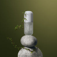 Load image into Gallery viewer, A serene, zen-inspired lifestyle shot of the Tonal Scenery Reparative Serum. The white bottle is balanced atop two smooth, stacked grey stones, surrounded by delicate green botanical sprigs against a soft olive-green background, reflecting the product's grounding and skin-strengthening qualities.