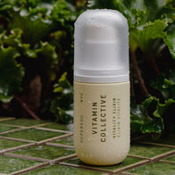 Load image into Gallery viewer, A lifestyle shot of the Vitamin Collective Vitality Elixir bottle resting on a green tiled surface in an outdoor garden setting. The bottle is covered in fresh water droplets, emphasizing its hydrating properties, with lush green foliage in the background.