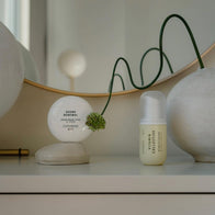 Load image into Gallery viewer, A minimalist lifestyle setup on a white vanity featuring the Vitamin Collective Vitality Elixir and the Sound Renewal Serum Cream. The products are arranged next to a large stone-like decorative object and a round mirror, reflecting a clean and modern aesthetic.